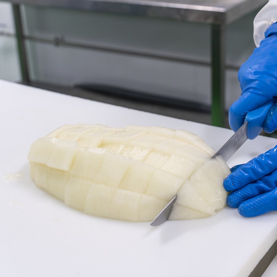 Manos enguantadas troceando melón sobre tabla de corte en la sala blanca